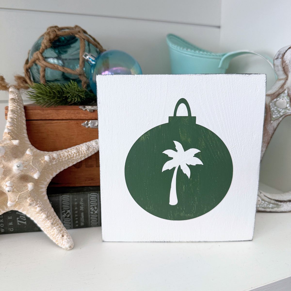 A square white decorative sign with a dark green Christmas ornament shape and a white palm tree silhouette, displayed on a shelf with nautical decor including a starfish and glass ornaments.
