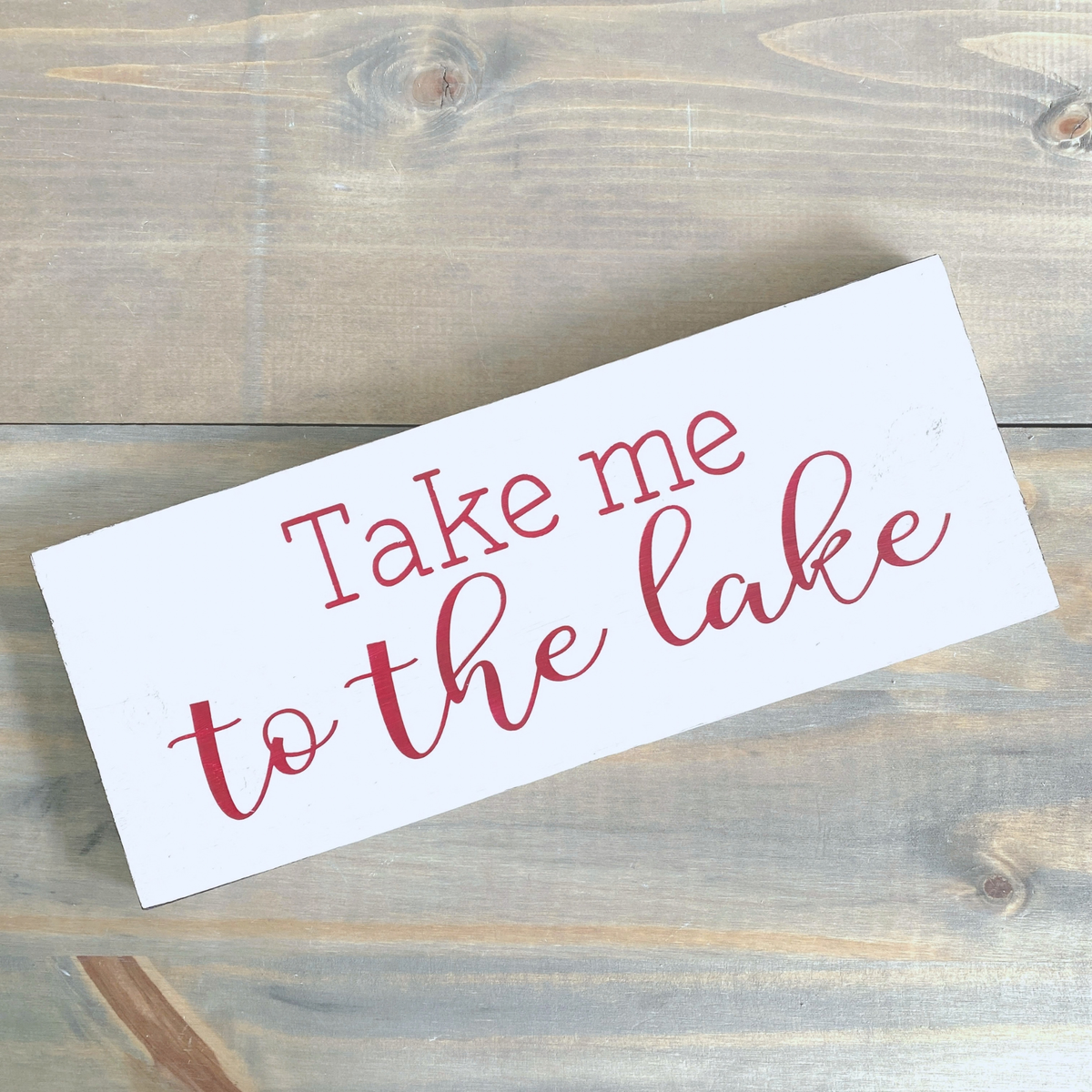 A white rectangular decorative sign with the phrase 'Take me to the lake' in red, photographed on a rustic wood plank surface.