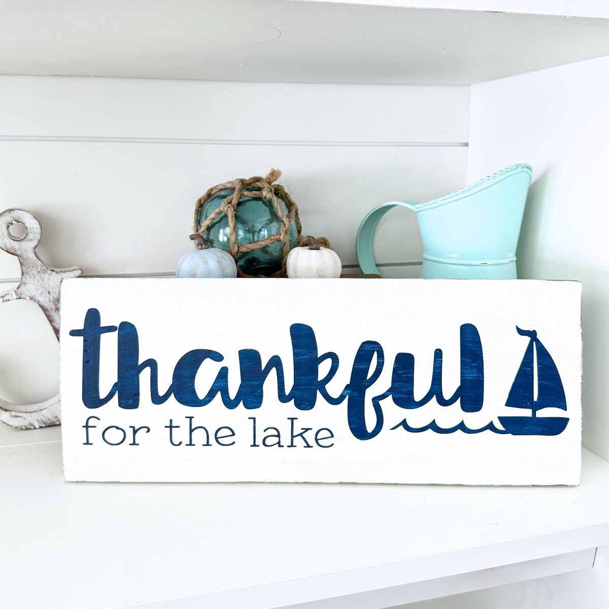 Thankful for the lake sign in white with navy lettering and sailboat and wave graphic. Placed on a white shelf with coastal and fall decor