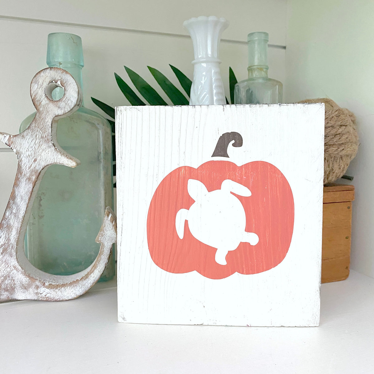 A square white wooden sign with a painted orange pumpkin and white turtle design, displayed on a shelf next to a distressed white anchor and other home decor items.