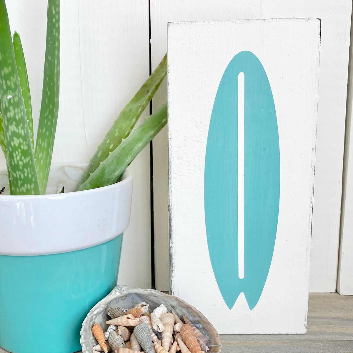 A white rectangular decorative sign with an aqua surfboard silhouette on the front, styled beside a potted aloe vera and a shell dish on a light wood surface.