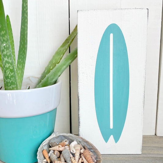 A white rectangular decorative sign with an aqua surfboard silhouette on the front, styled beside a potted aloe vera and a shell dish on a light wood surface.