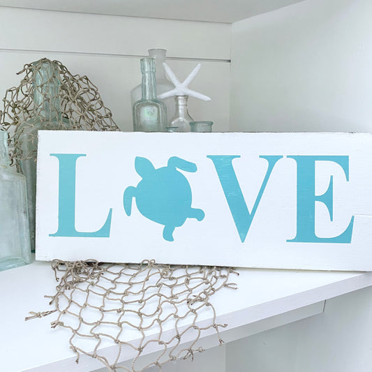 A white rectangular decorative sign with the word LOVE, where the O is replaced by an aqua sea turtle silhouette. The sign is styled on a white shelf with nautical decor, including glass bottles, a starfish, and netting.