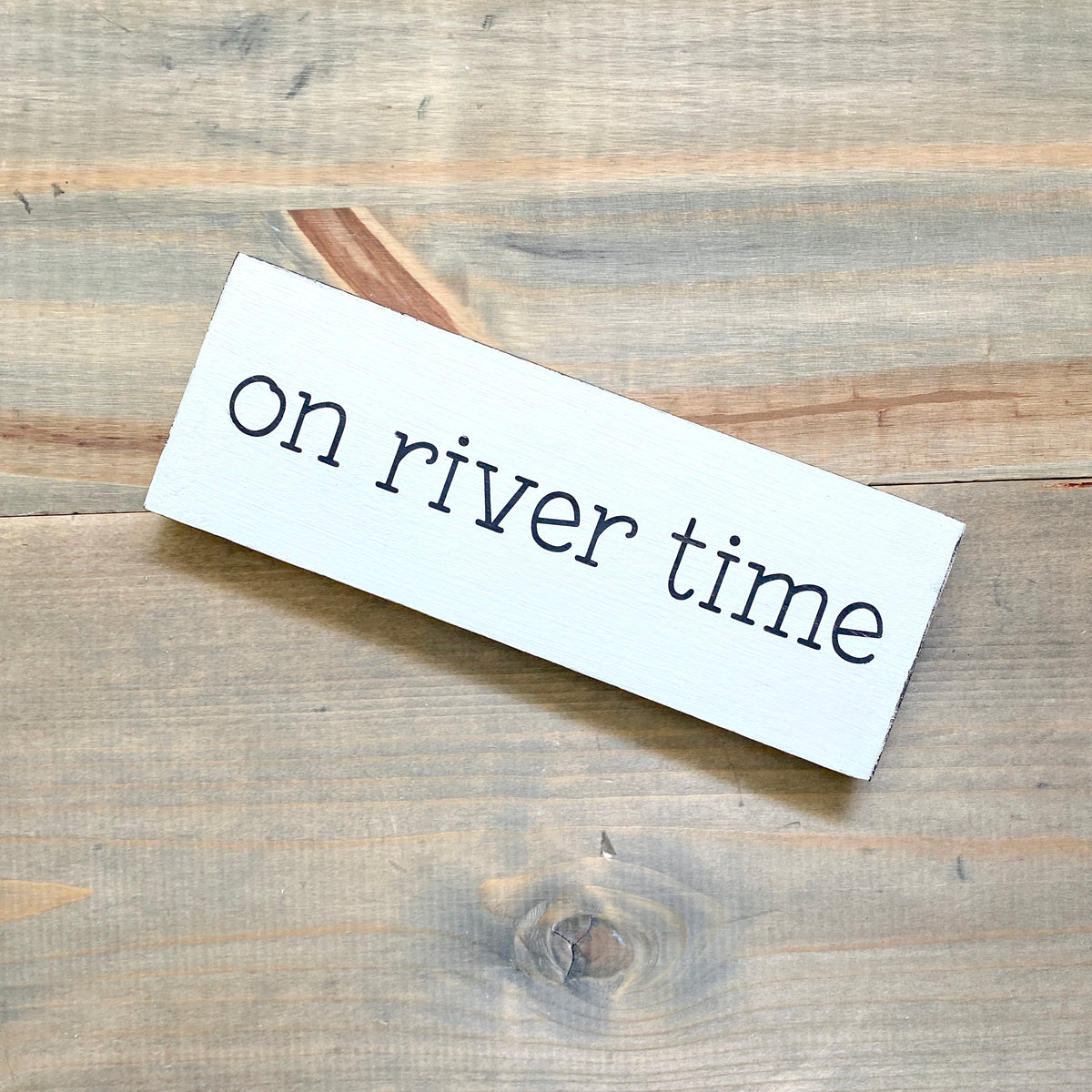 A white rectangular sign with the phrase 'on river time' in black lettering, photographed on a rustic light brown wood plank surface.