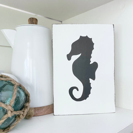 A white rectangular decorative sign with a large black seahorse silhouette, displayed on a white shelf next to a white ceramic pitcher and a blue glass ornament.