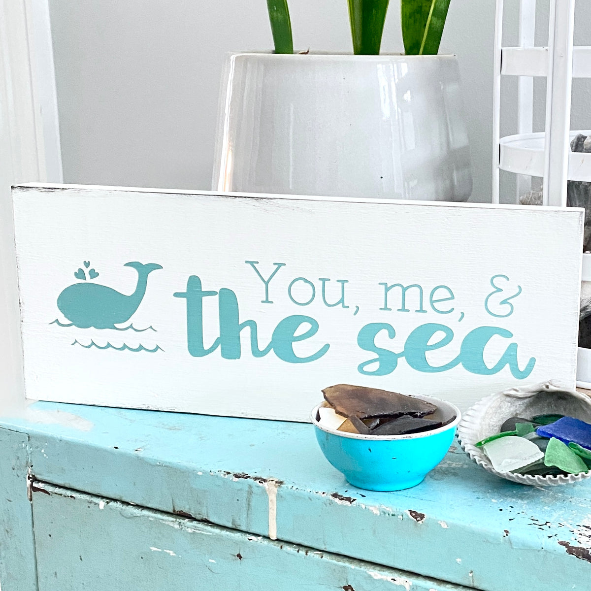 A white wooden sign with a distressed finish featuring the text 'You, me, & the sea' in aqua lettering and an aqua whale illustration. The sign is styled on a distressed light blue metal cabinet with small decorative bowls and a white ceramic planter in the background
