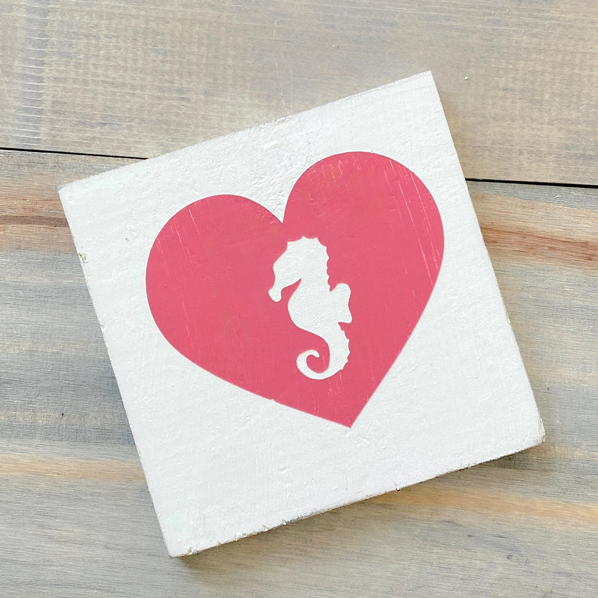 A square white painted wooden sign featuring a large pink heart with a white seahorse silhouette in the center, photographed on a rustic wood surface.