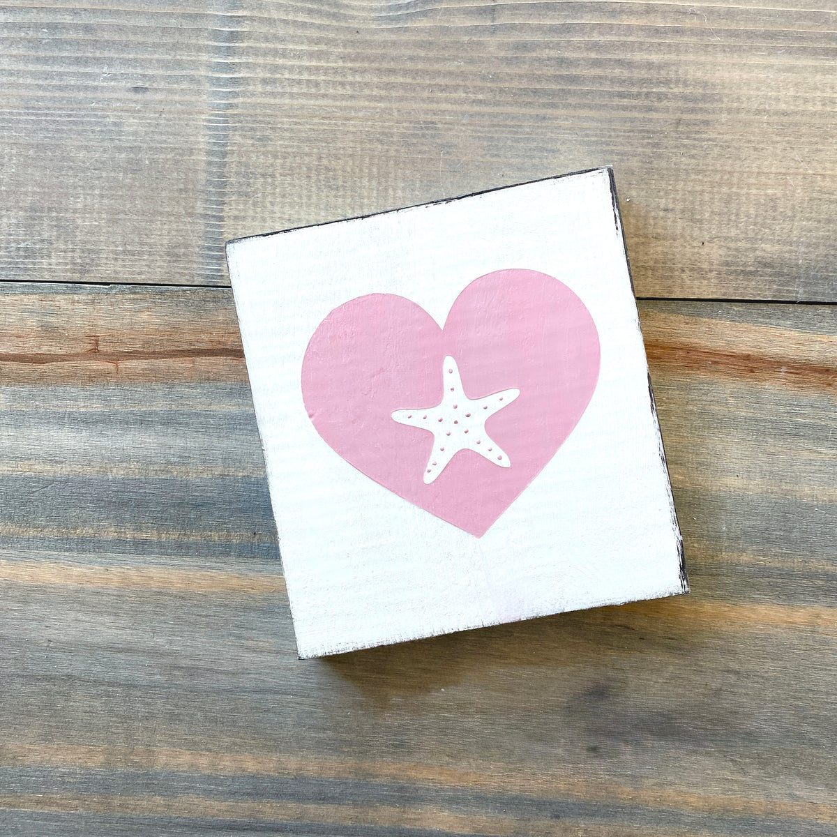 A square white decorative sign with a pink heart and white starfish design, photographed on a rustic wood plank surface.