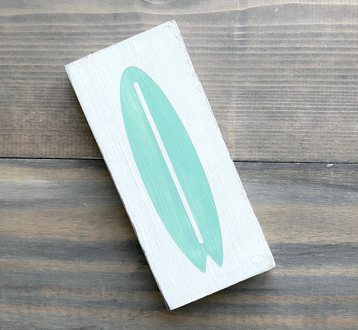 A rectangular white wooden sign featuring a painted mint green surfboard design, placed diagonally on a dark wood plank surface.