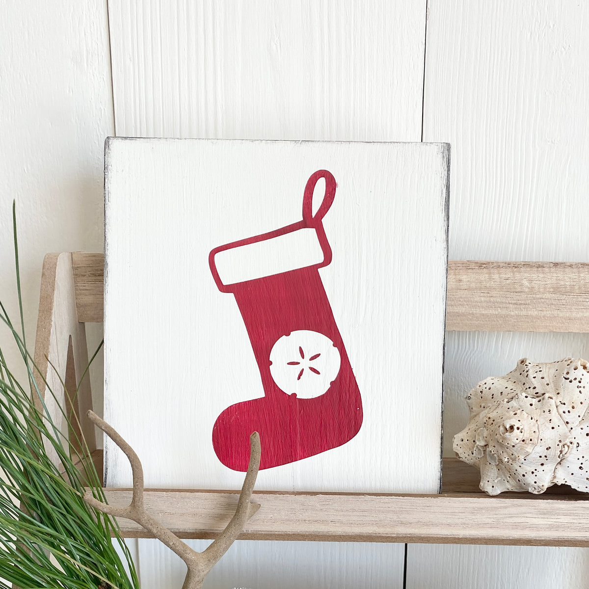 A square white decorative sign with a red Christmas stocking with a sand dollar  painted on it, displayed on a light wood shelf with a coral shell and green plant leaves nearby.