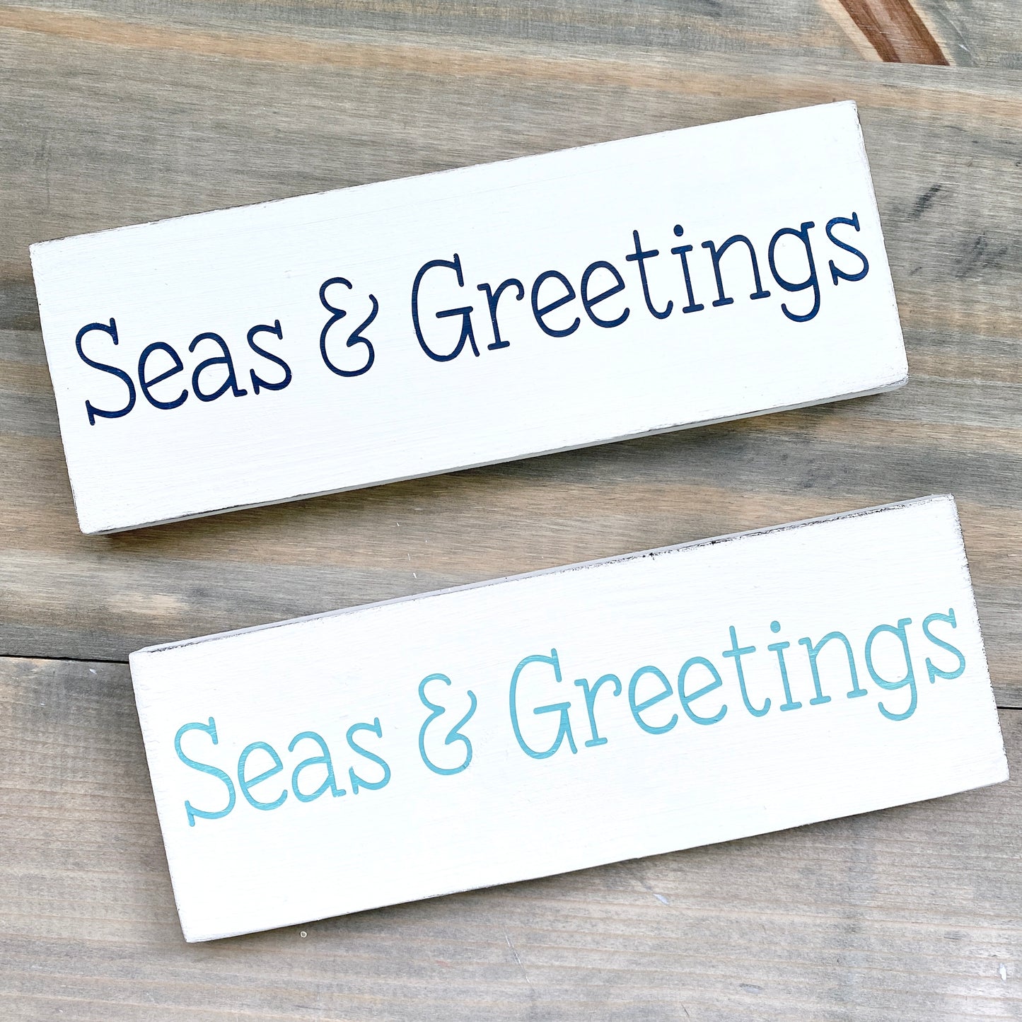 Two rectangular white wooden signs with the phrase "Seas & Greetings" one in dark navy blue and the other in aqua displayed on a rustic gray wood background.