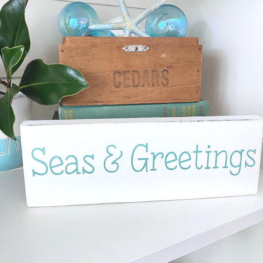 A white rectangular wooden sign with the words 'Seas & Greetings' in aqua, displayed on a white shelf in a nautical-themed setting with a wooden crate, books, starfish, and blue glass orbs.