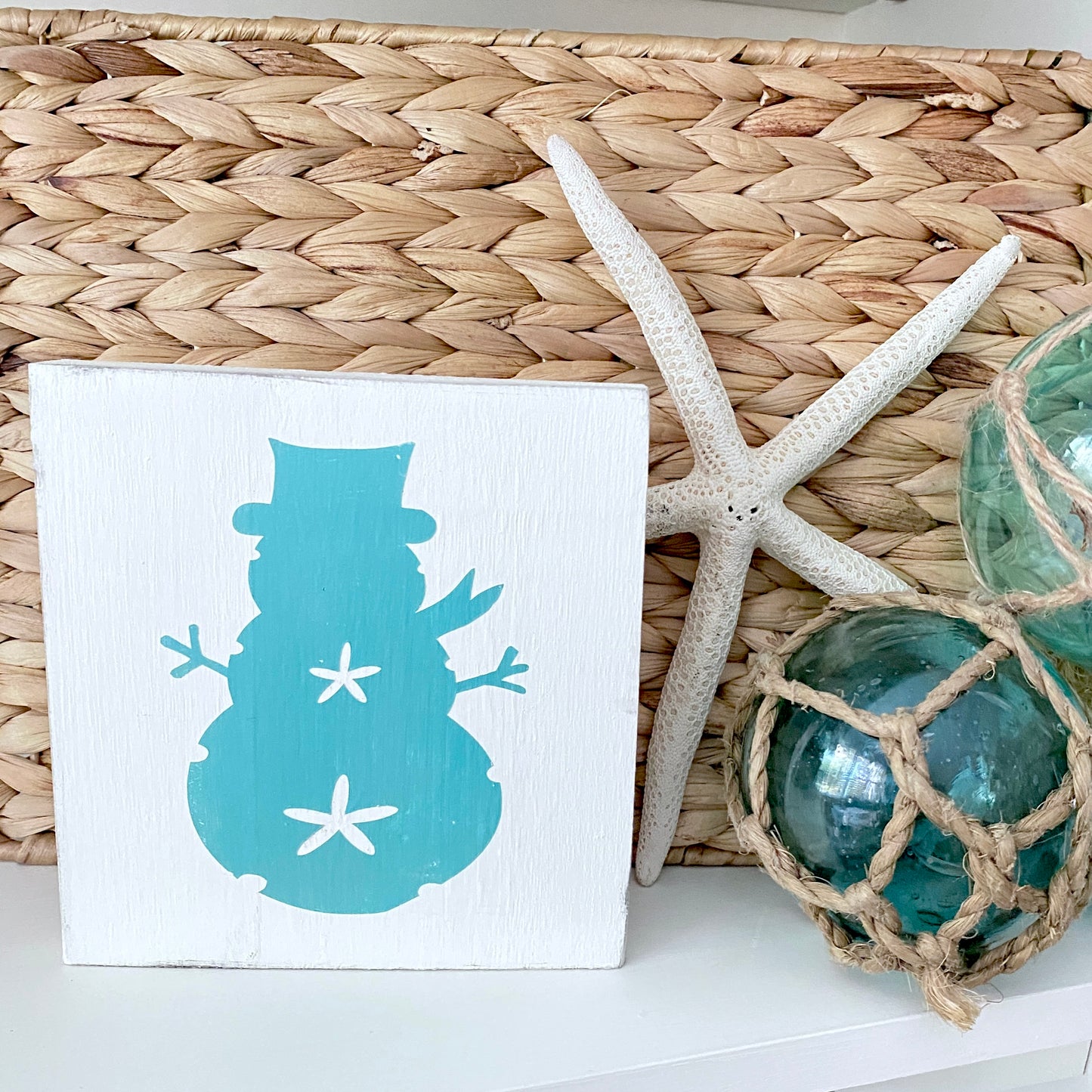 A square white decorative sign featuring a aqua snowman silhouette with white starfish accents, styled on a white surface next to a woven basket, a white starfish, and green glass orbs.