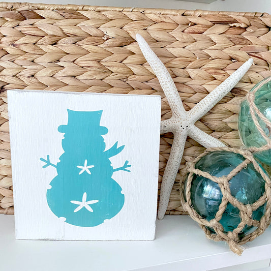 A square white decorative sign featuring a aqua snowman silhouette with white starfish accents, styled on a white surface next to a woven basket, a white starfish, and green glass orbs.