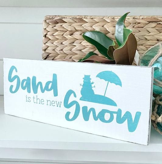 A rectangular white wooden sign with the phrase 'Sand is the new Snow' in light blue lettering, featuring a blue snowman under an umbrella graphic. The sign is styled in a home setting with a woven basket and green leaves in the background.