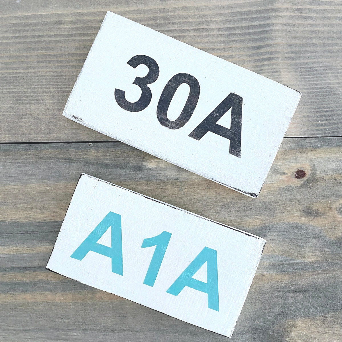 Two white rectangular signs with black and aqua lettering, reading '30A' and 'A1A' respectively, placed on a rustic wooden surface.