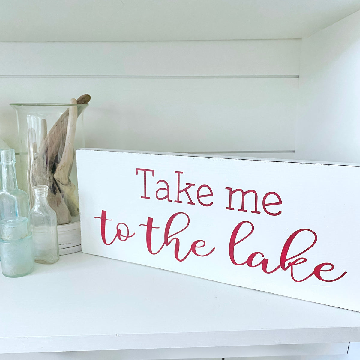 A white rectangular wooden sign with the phrase 'Take me to the lake' in red script, displayed on a white shelf next to glass bottles and driftwood.