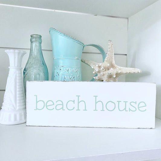 A white rectangular sign with the words 'beach house' in a pale mint lowercase font, styled on a white shelf with a turquoise pitcher, a starfish, a glass bottle, and a white vase.