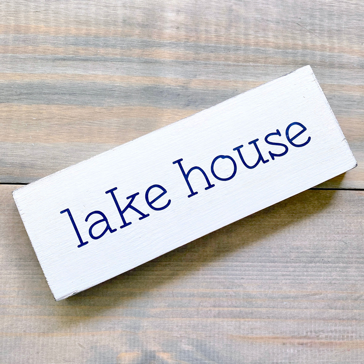 A rectangular white wooden sign with the words 'lake house' in navy, photographed on a rustic light brown wood surface.