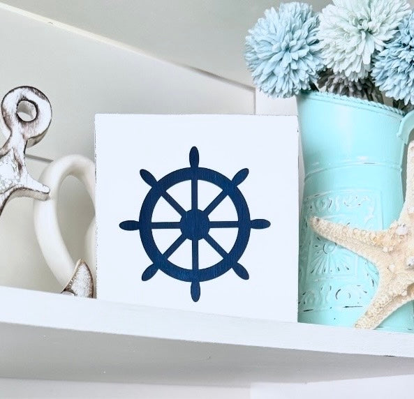 A square white decorative sign featuring a navy blue ship's wheel motif, styled on a white shelf with a light blue flower pot, and a starfish.