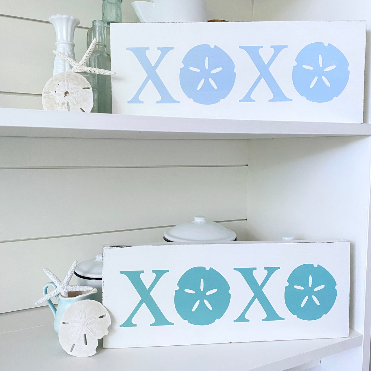 Two white decorative wooden signs with sandal-shaped letter cutouts, displayed on white shelves. The top sign reads 'XOXO' in light blue, and the bottom sign reads 'XOXO' in aqua.
