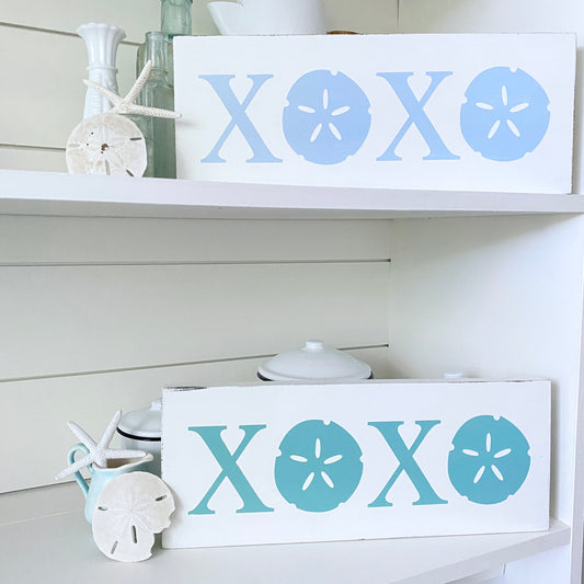 Two white decorative wooden signs with sandal-shaped letter cutouts, displayed on white shelves. The top sign reads 'XOXO' in light blue, and the bottom sign reads 'XOXO' in aqua.
