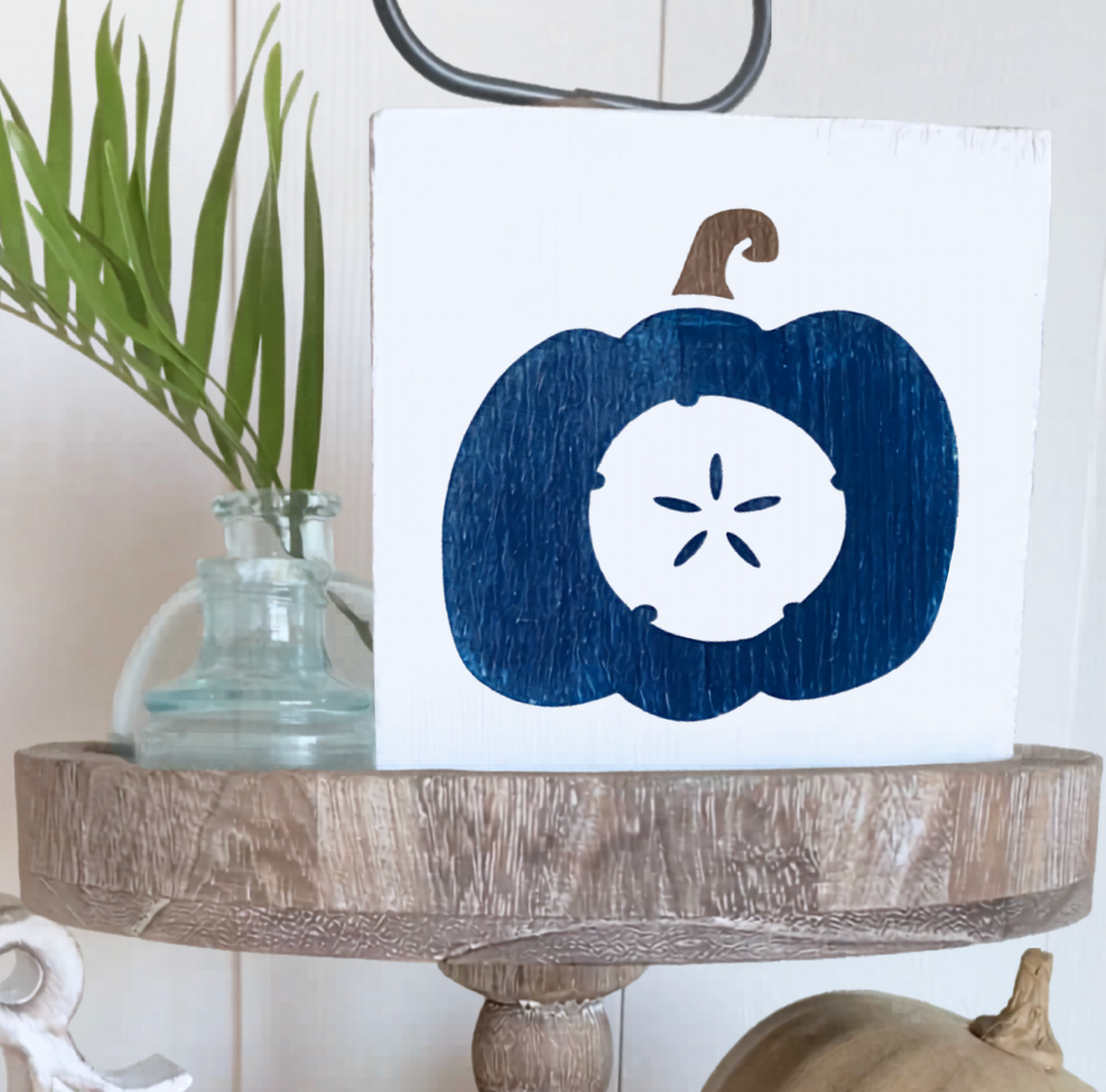 A square white decorative sign featuring a navy blue pumpkin silhouette with a white sand dollar cutout in the center, styled on a rustic wooden stand next to a small glass vase with green leaves.