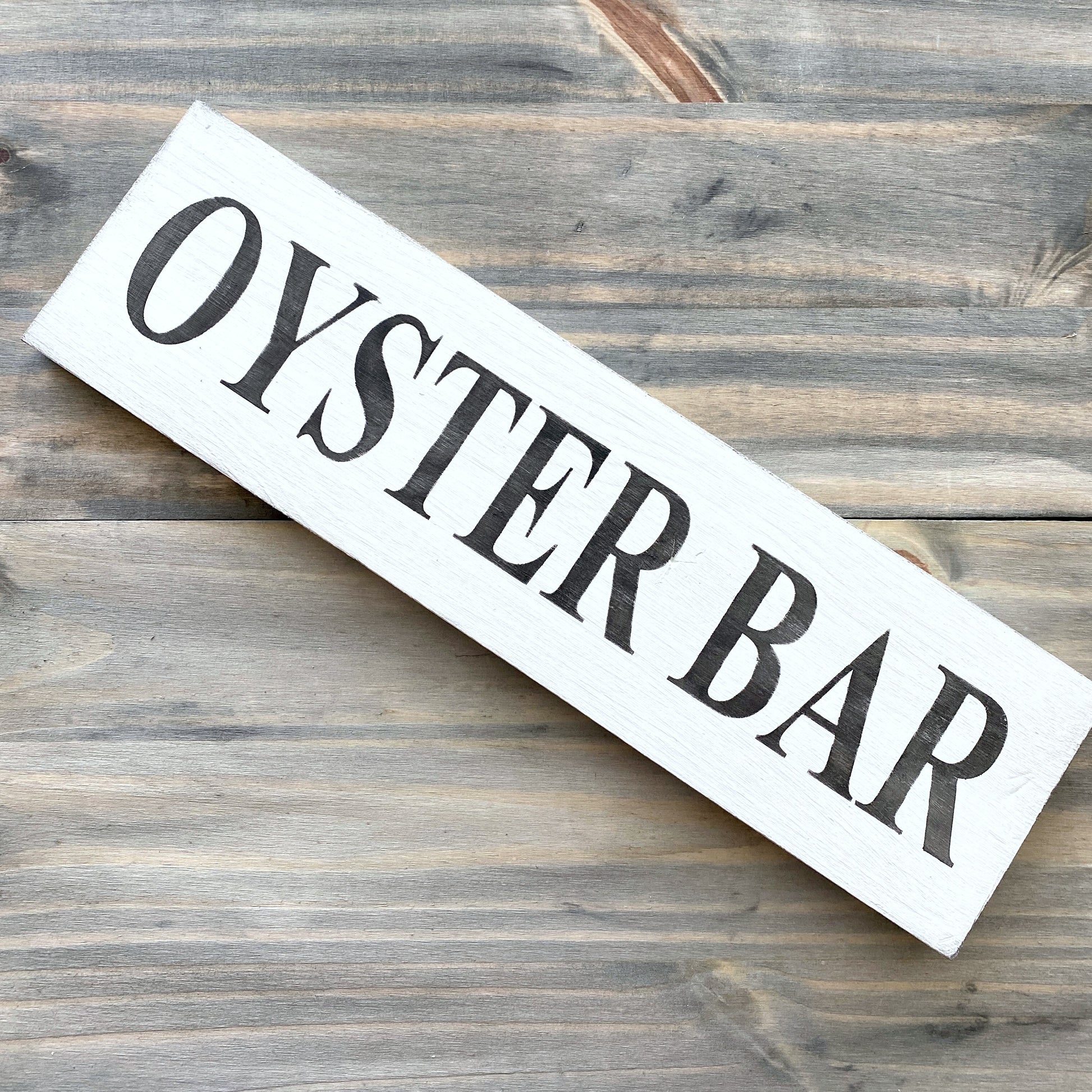 A rustic white wooden sign with black text reading 'OYSTER BAR', placed diagonally on a weathered gray-brown wooden plank surface.