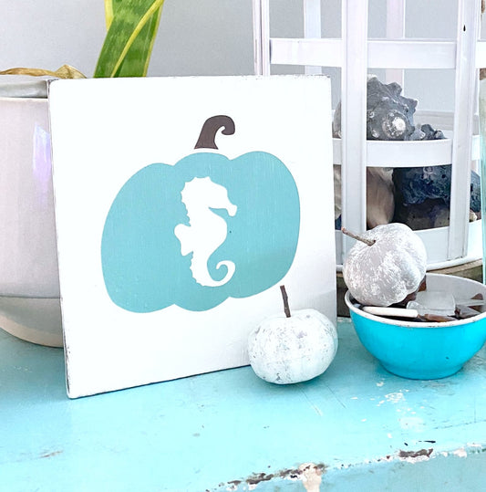 A square white decorative sign featuring an aqua pumpkin silhouette with a white seahorse, styled on a turquoise surface with small pumpkins and a bowl of sea glass and other coastal decor nearby.