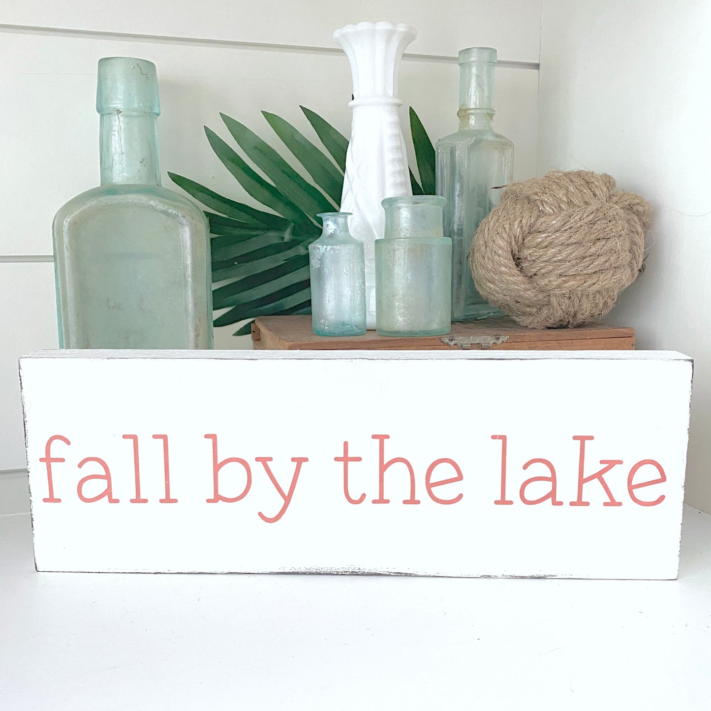 Decorative shelf with bottles, a plant, and a ball of rope on a white surface with 'fall by the lake' sign.