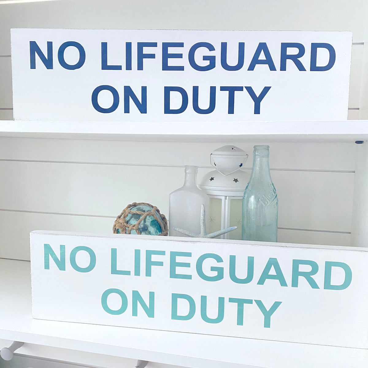 Two white wooden signs with the text 'NO LIFEGUARD ON DUTY' displayed on white shelves. The top sign has dark blue lettering and the bottom sign has light aqua lettering.