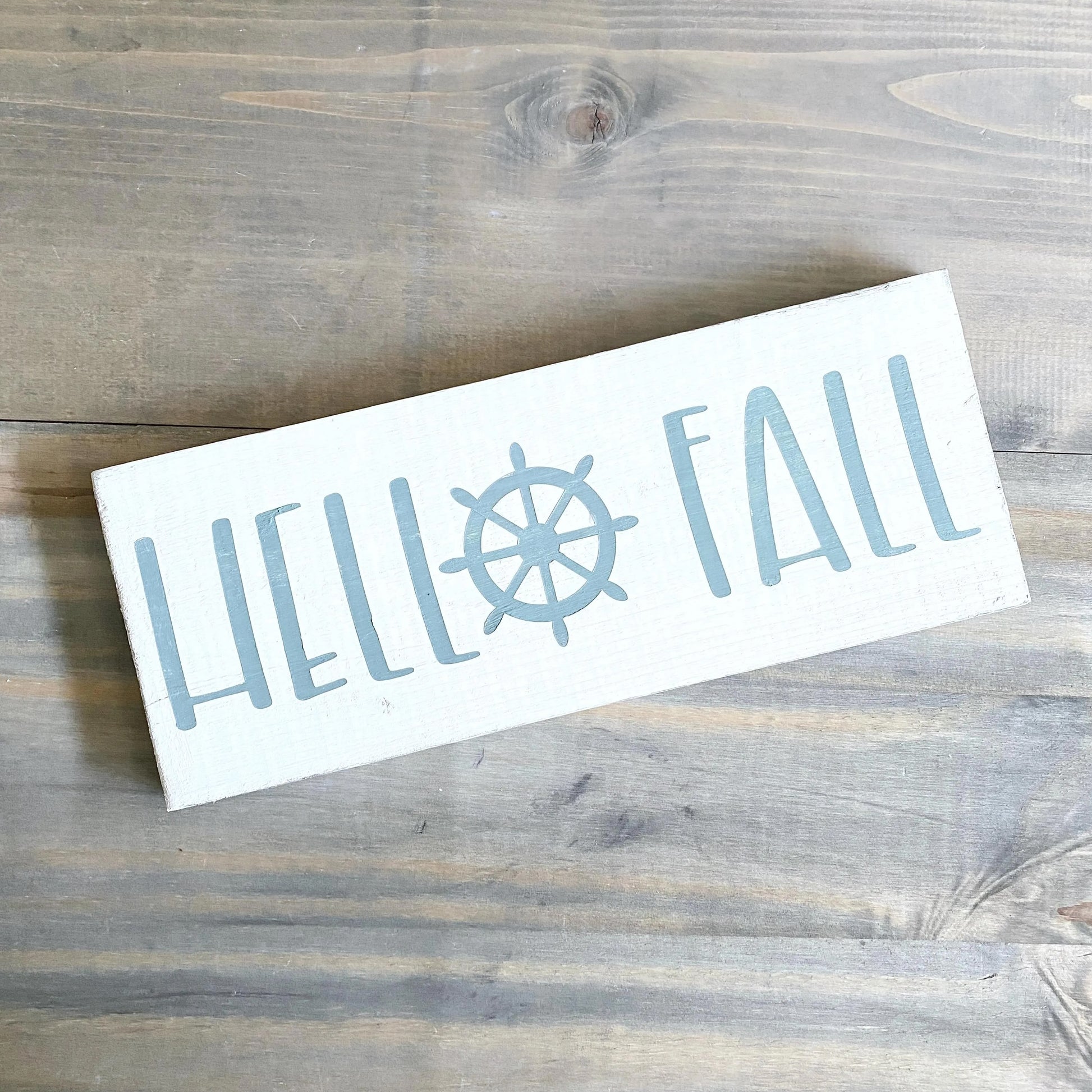 Rectangular sign with 'HELLO FALL' text and a ship's wheel on a wooden surface