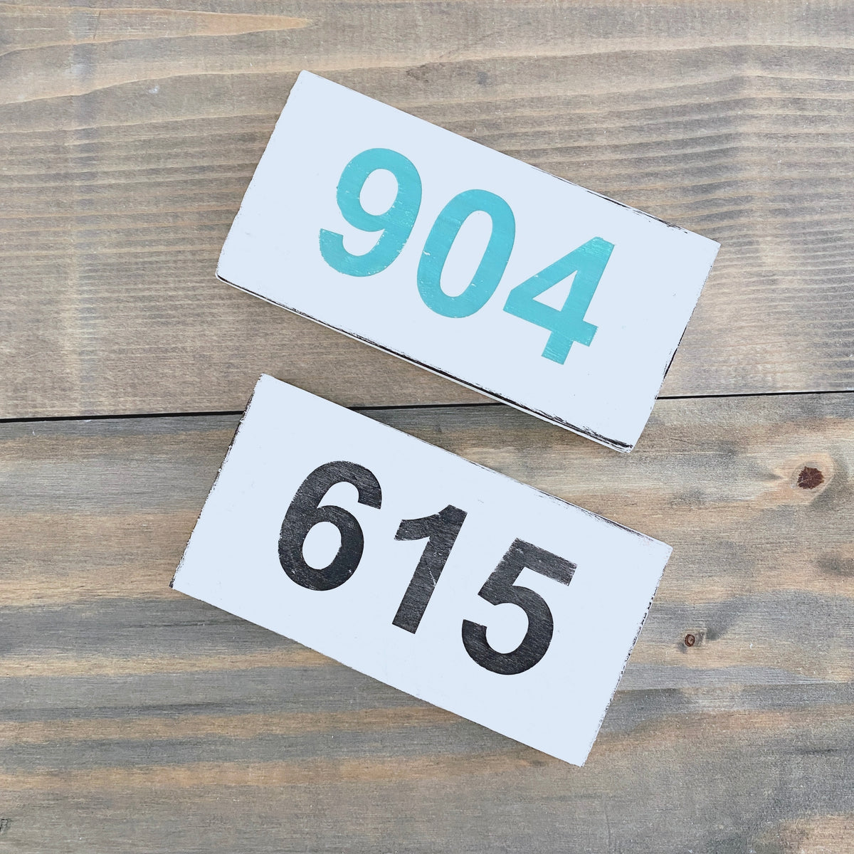 Two rectangular white house number signs with rustic edges, one displaying '904' in aqua and the other '615' in black, arranged diagonally on a weathered wood plank surface.