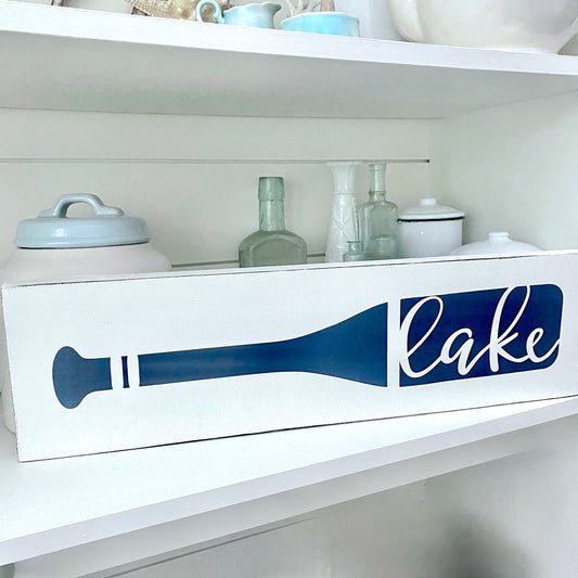 A white decorative sign with a navy blue oar silhouette and the word "lake" in white script, displayed on a white shelf with glass bottles and ceramic containers behind it.