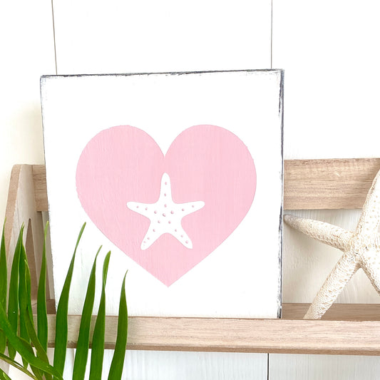 A square white wall sign with a distressed border, featuring a pink heart and a white starfish design. The sign is styled on a light wood shelf next to a real starfish and green plant leaves against a white paneled wall.