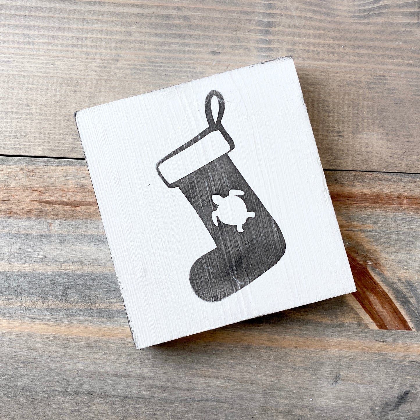 A square white wooden sign with a black painted Christmas stocking and a small white turtle design, photographed on a rustic wood plank surface.