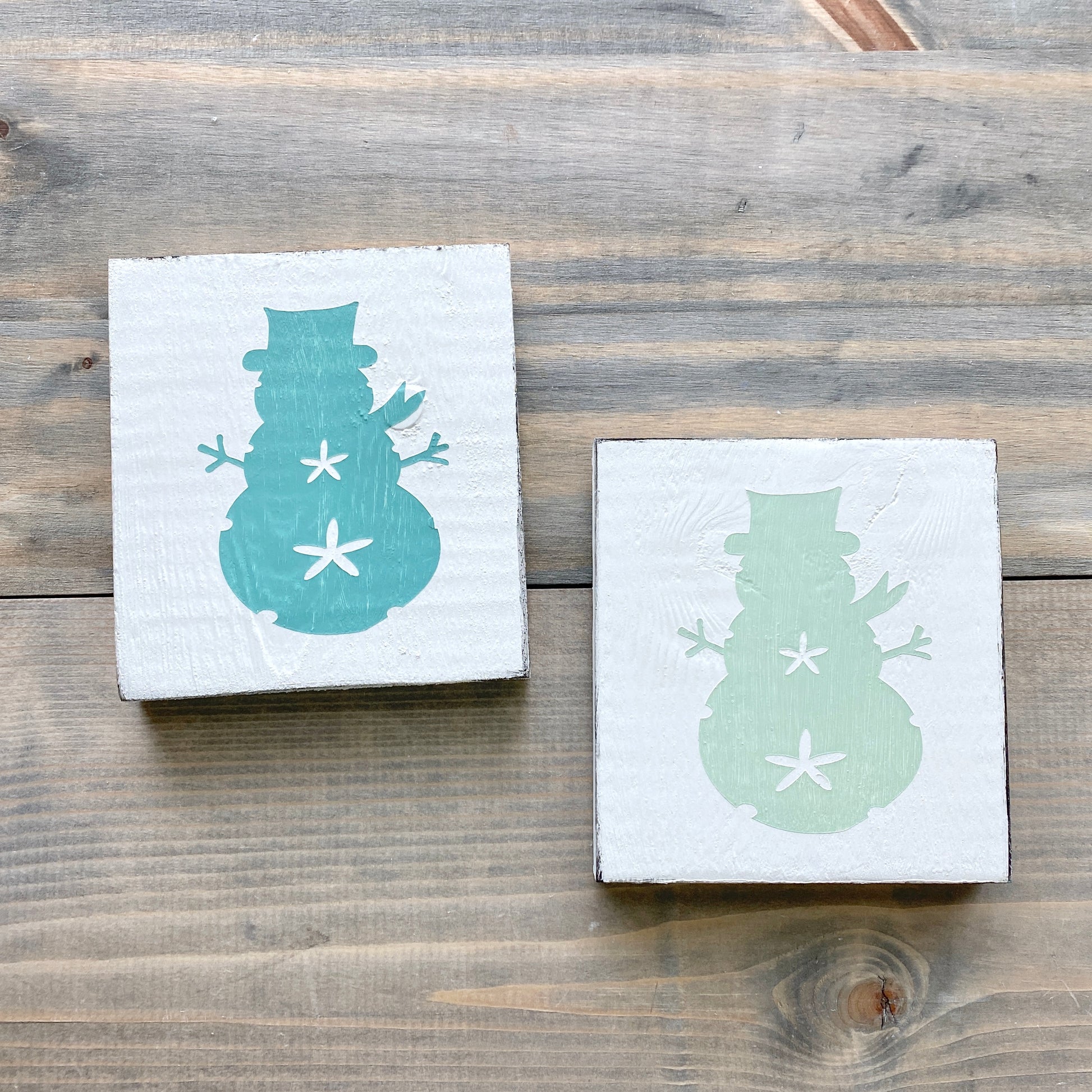 Two square white wooden signs with painted snowman designs, one in aqua and one in mint displayed on a rustic gray-brown wooden surface.