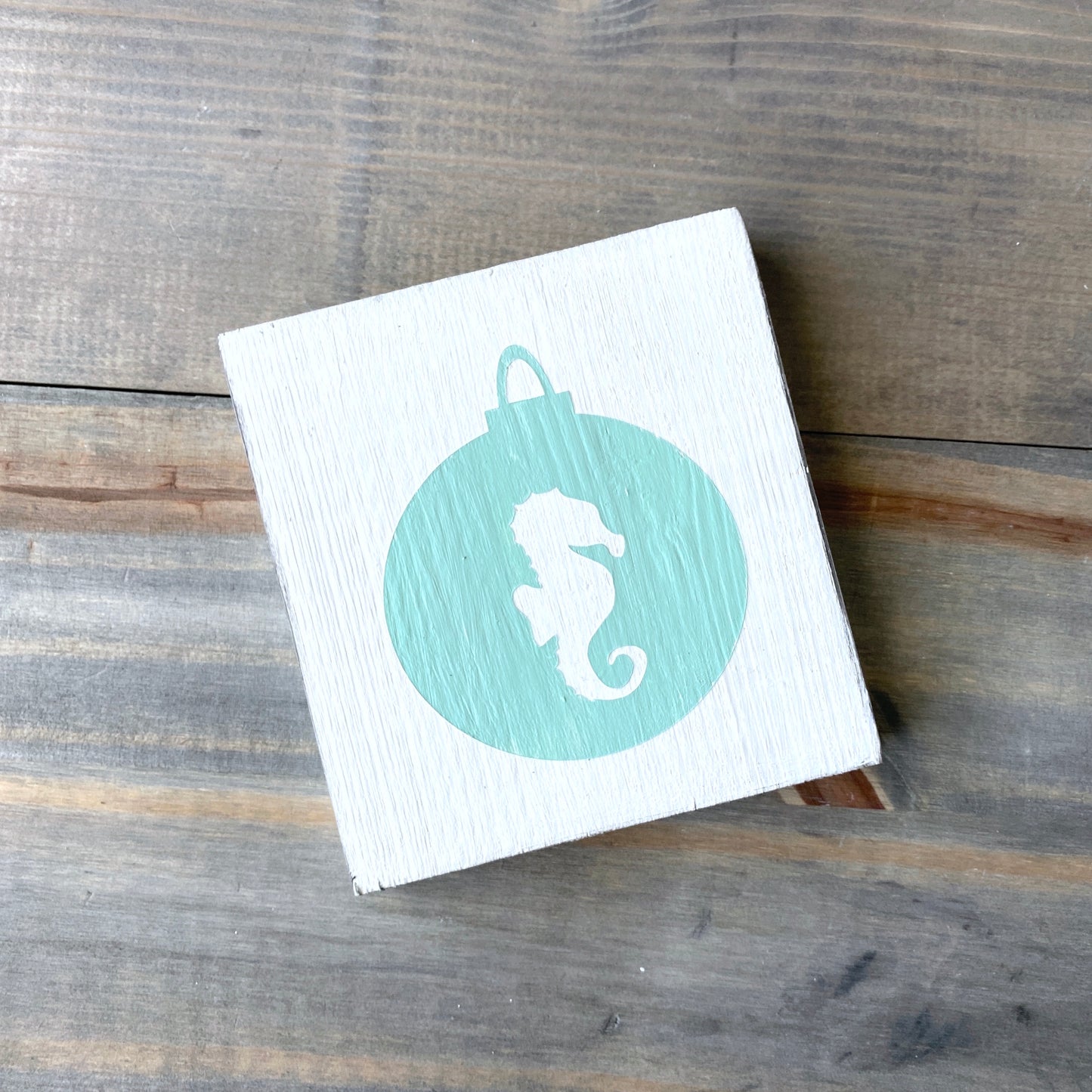 A square white wooden plaque featuring a mint ornament design with a white seahorse silhouette, displayed on a rustic gray-brown wooden surface.