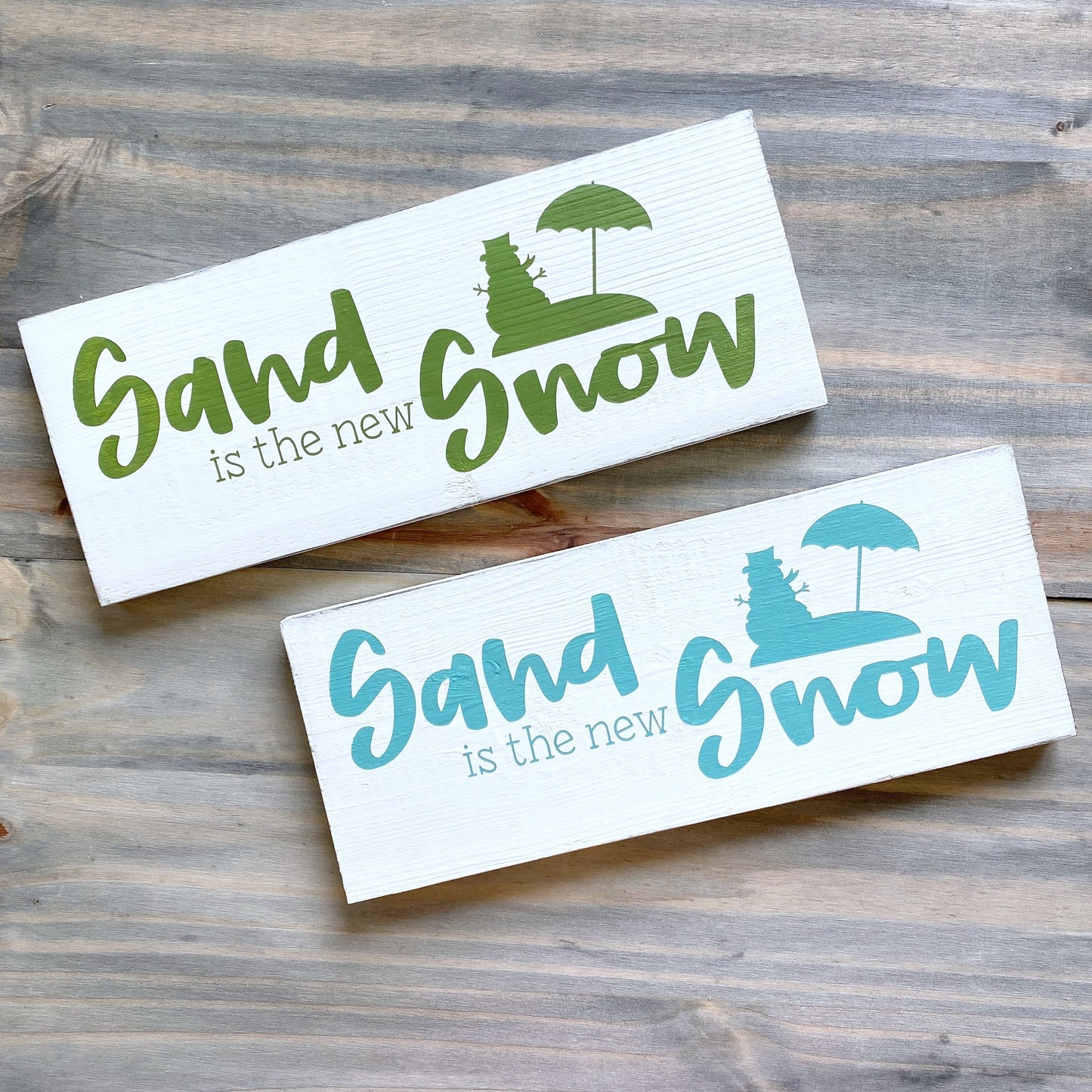 Two white rectangular decorative signs with the phrase 'Sand is the new Snow' in blue and green lettering, plus a small beach umbrella and tree graphic, displayed on a rustic wood surface.