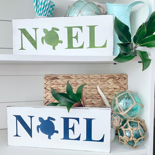Two white wooden 'NOEL' sign boards with turtle cutouts, styled on white shelves with decorative items including a woven basket, blue glass ornaments, green leaves, and a light blue pitcher.