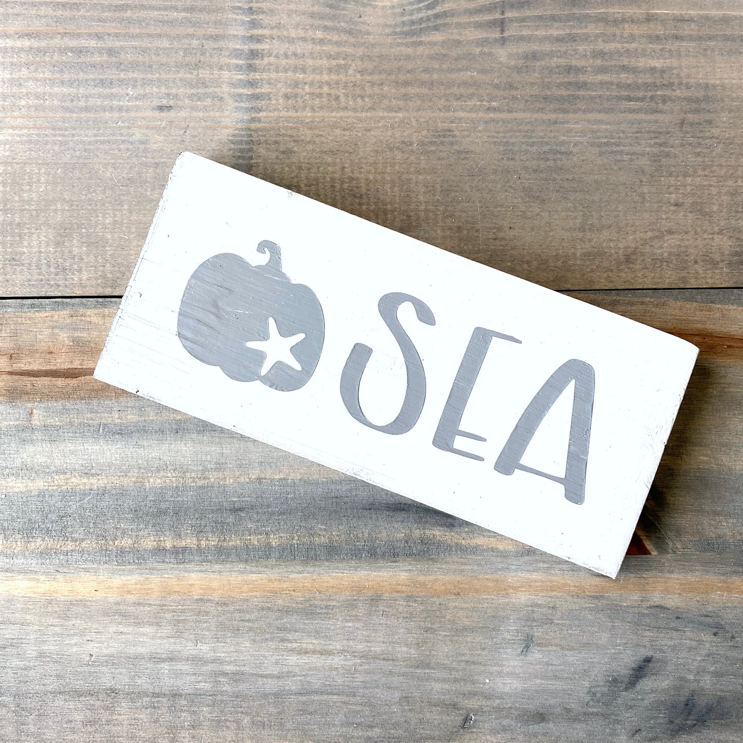 A rectangular white wooden sign with a gray painted pumpkin and starfish and the word 'SEA' in large gray letters, placed diagonally on a rustic wood plank surface.