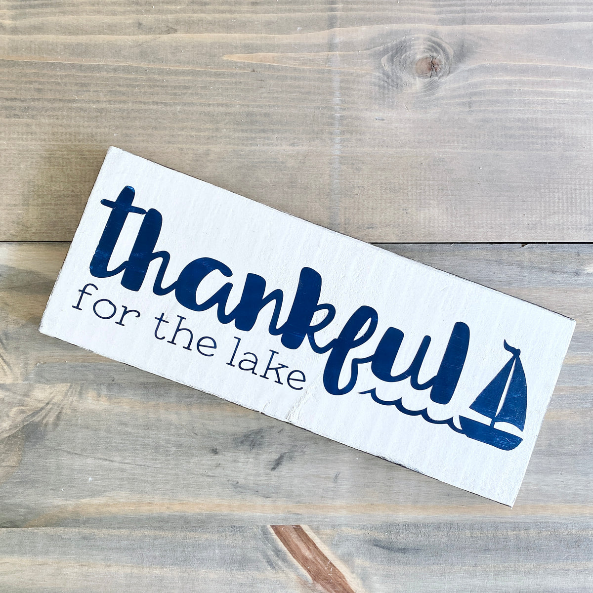 Thankful for the lake sign in white with navy lettering and sailboat and wave graphic. Laying on a rustic wood background