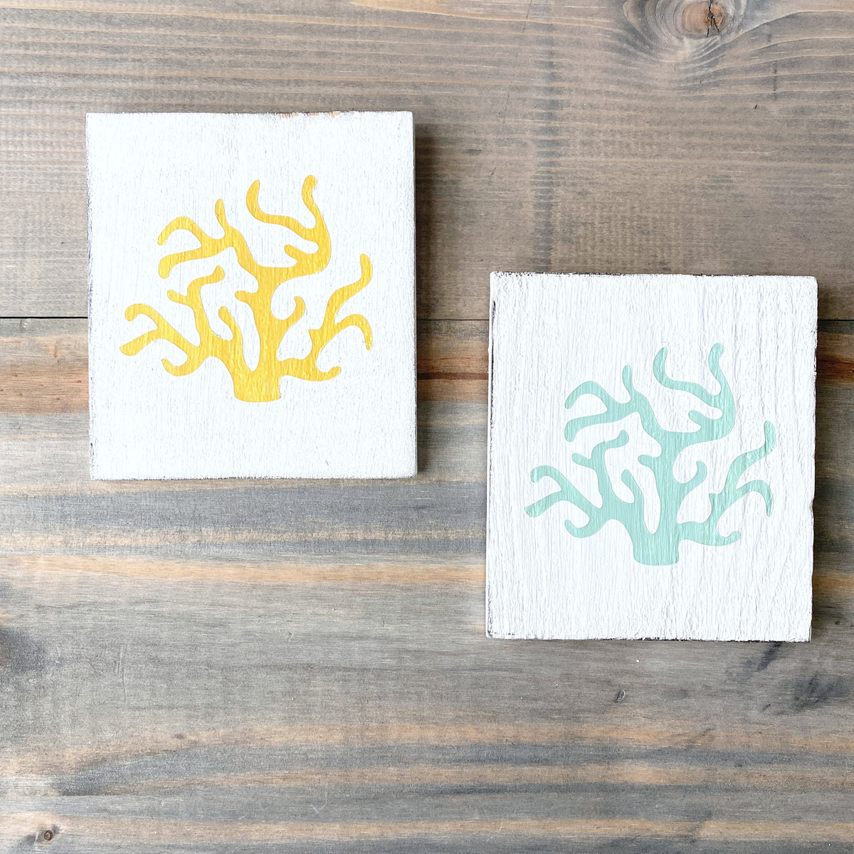 Two square white wooden signs with coral designs, one in yellow and one in mint, displayed on a rustic wooden surface.