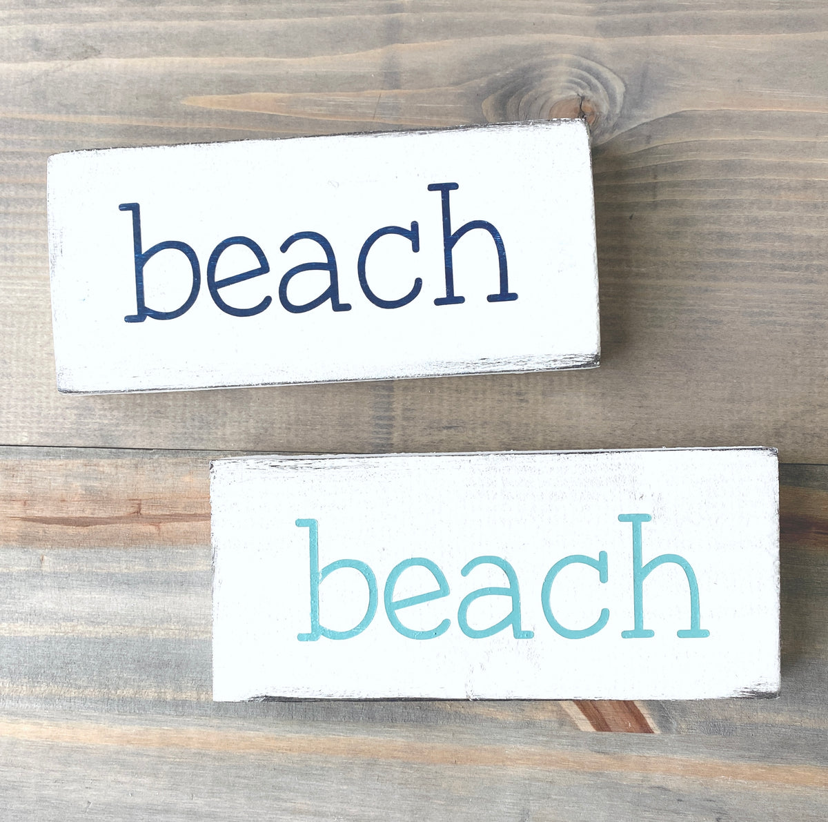 Two rectangular white wooden signs with the word "beach" printed in lowercase letters, one in dark navy and one in aqua, displayed on a rustic gray-brown wooden surface.