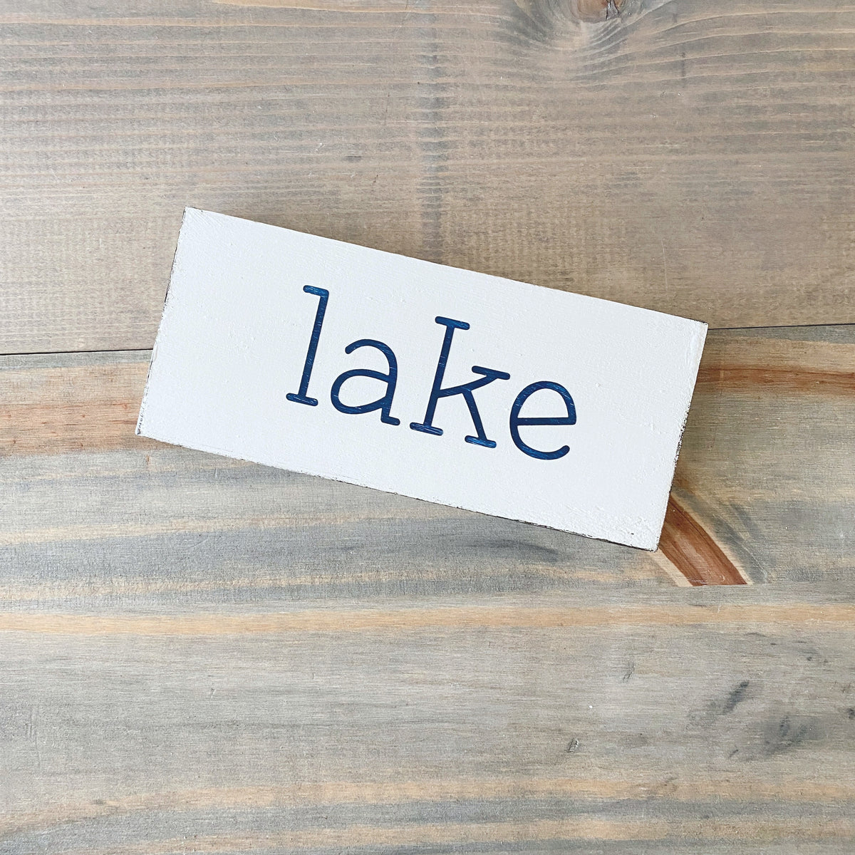 A white rectangular wooden sign with the word 'lake' written in dark blue lowercase serif letters, placed on a rustic gray-brown wood plank surface.
