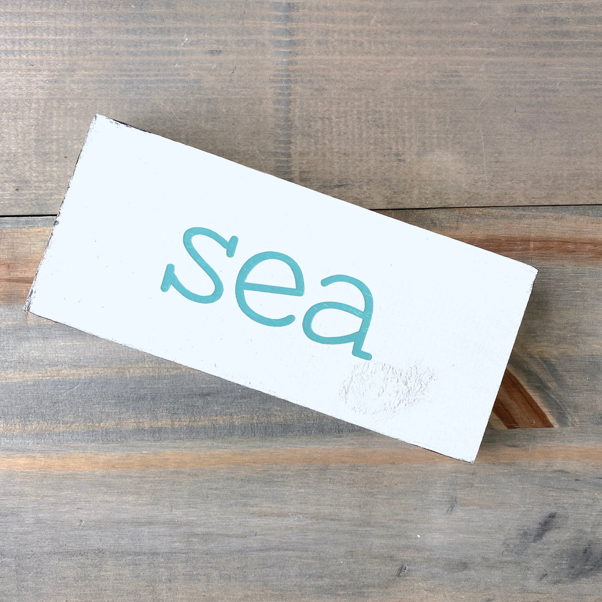 A white rectangular wooden sign with the word 'sea' painted in aqua lowercase letters, photographed on a rustic gray-brown wooden surface.
