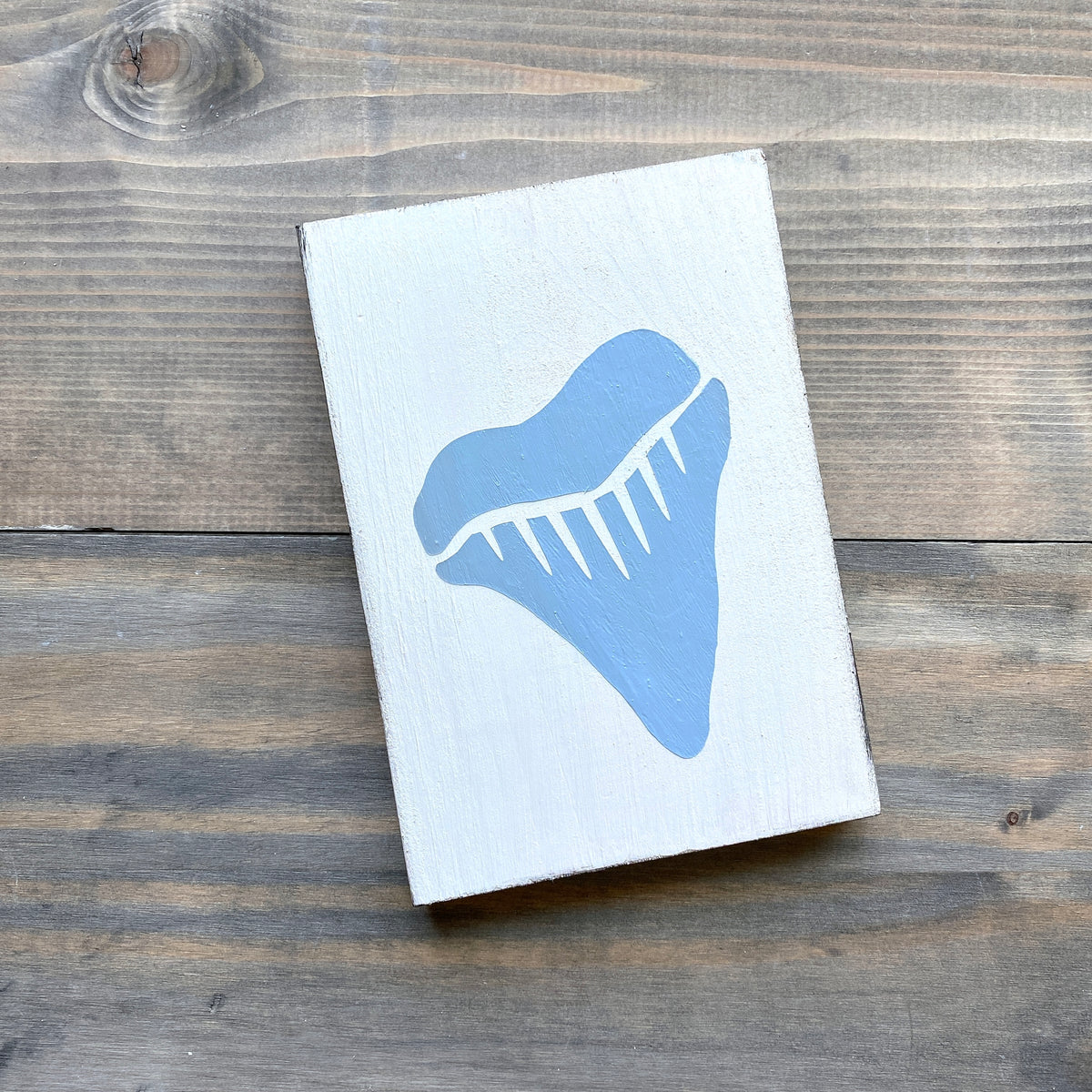 A rectangular white wooden block or sign featuring a large light blue shark tooth graphic, photographed from above on a rustic dark wood plank surface.
