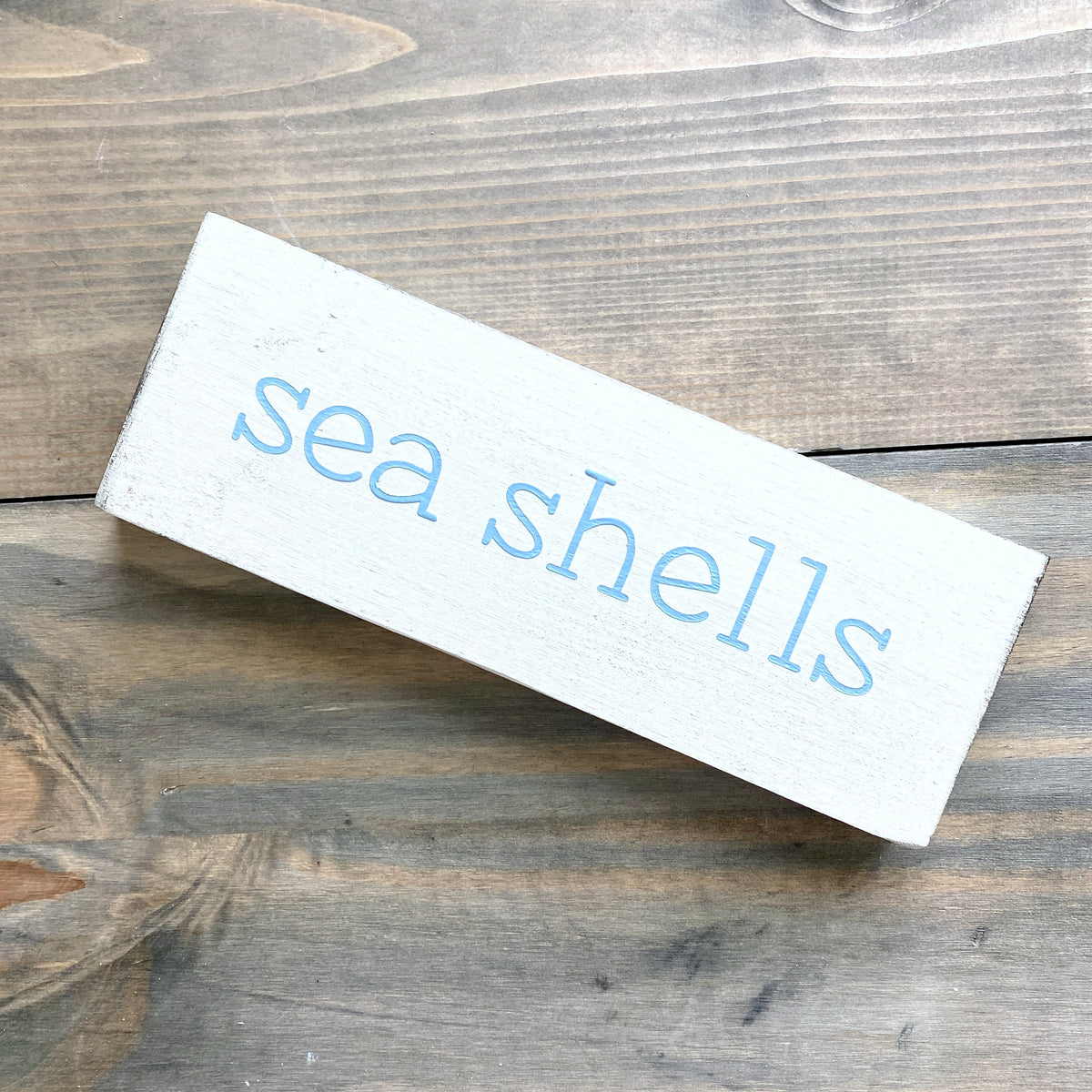 A rectangular white wooden sign with the words 'sea shells' painted in light blue lowercase letters, resting on a rustic brown wooden surface.