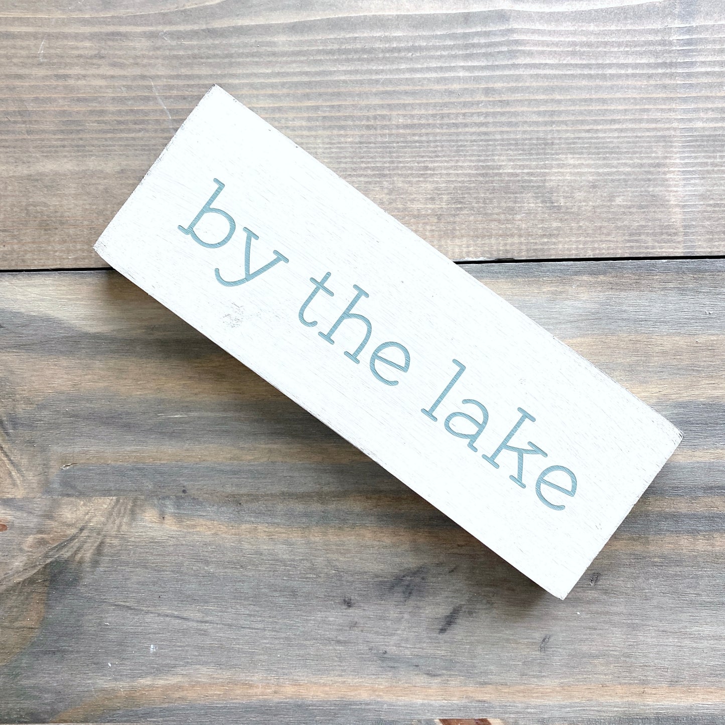 Rectangular wooden sign with 'by the lake' text on a wooden surface