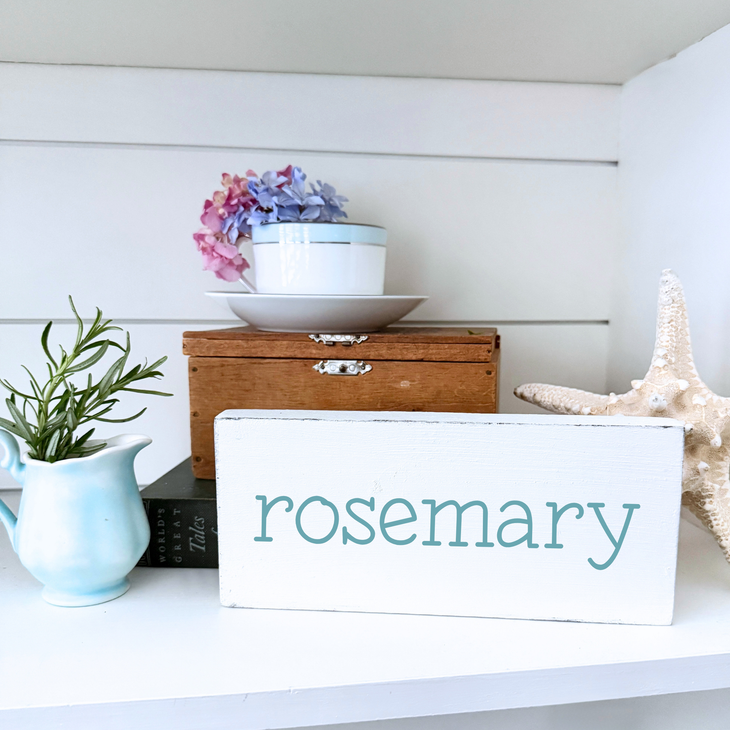 White sign labeled 'rosemary' on a shelf with decorative items including a starfish and a rosemary plant.